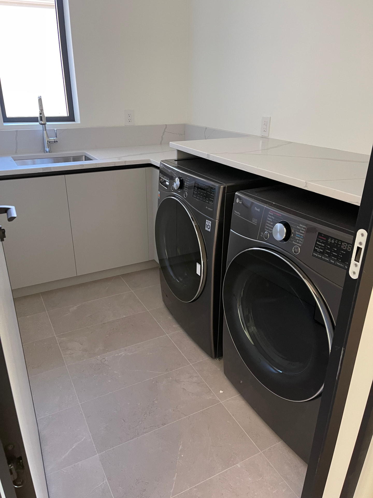 Light grey flat-panel laundry cabinets with black channel pulls, white marble-veined quartz counters, a folding counter over a graphite front-load washer and dryer, and an undermount sink with a gooseneck faucet