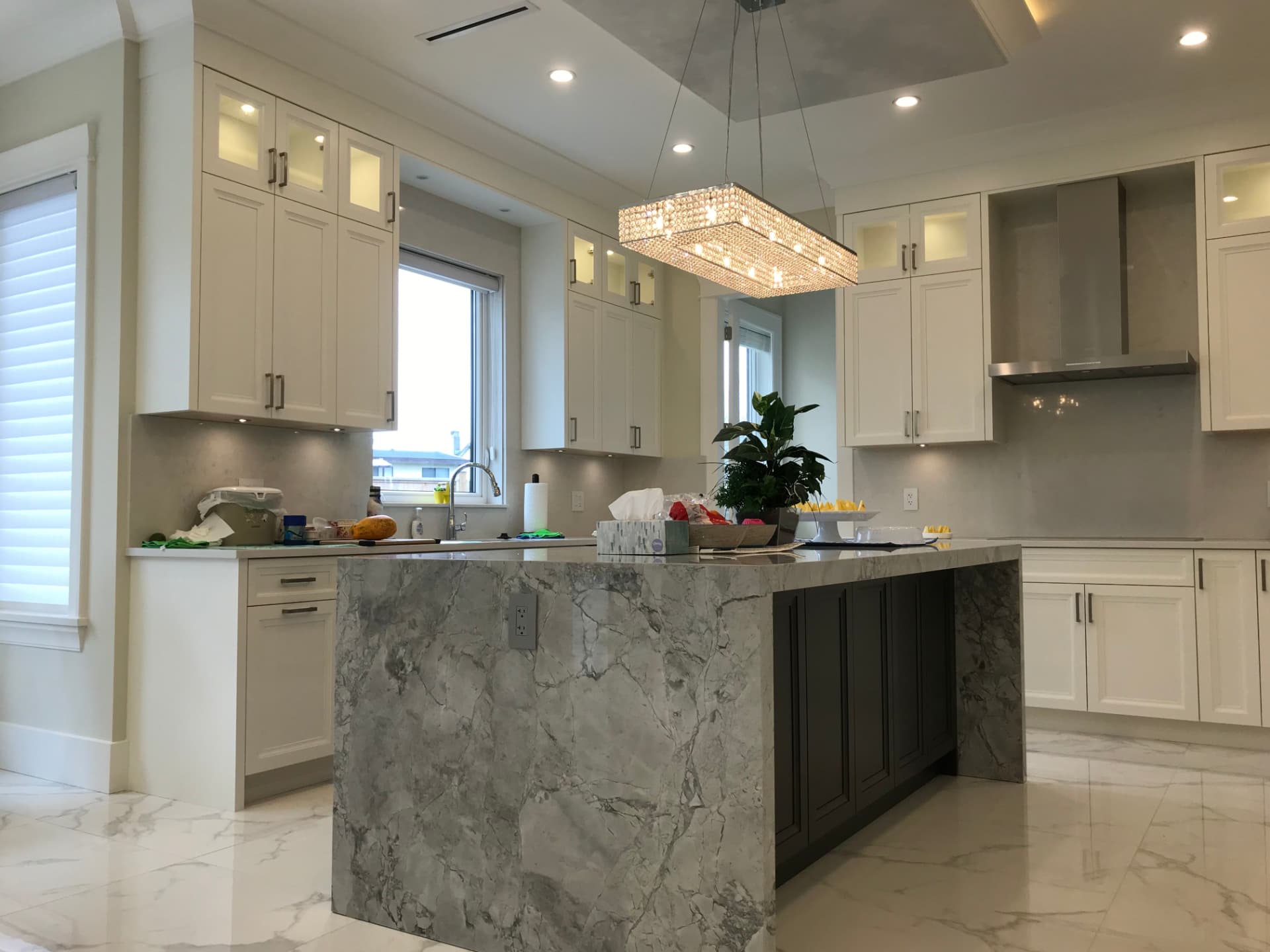 Cream shaker kitchen with a charcoal-black island under a grey-veined marble waterfall top, white quartz counters and slab backsplash, glass-front display uppers, built-in ovens, and a crystal chandelier