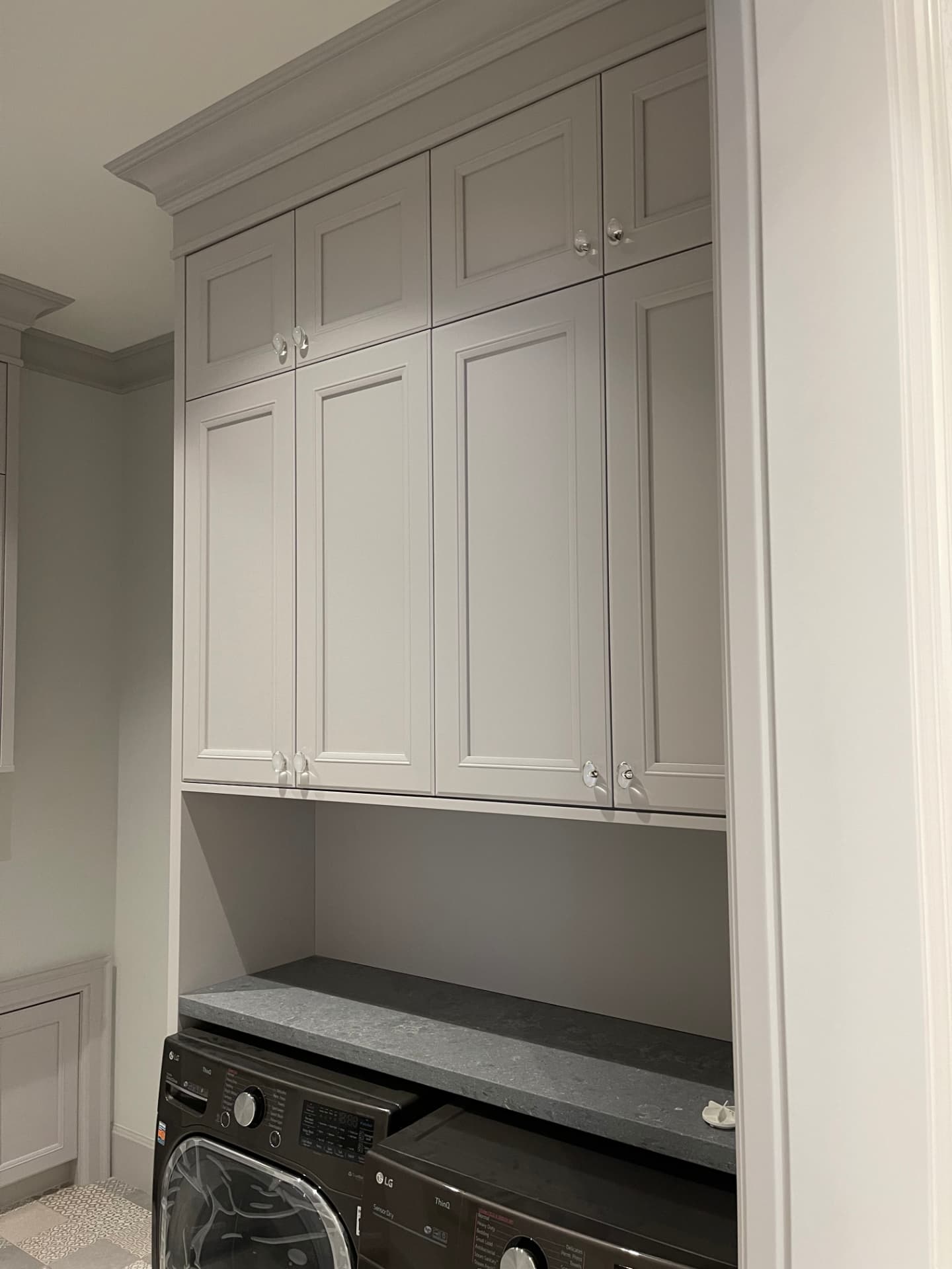 Pale greige laundry room cabinets with crown molding and crystal knobs, dark stone counters over a washer and dryer, and a sink area with an undermount sink and gooseneck faucet