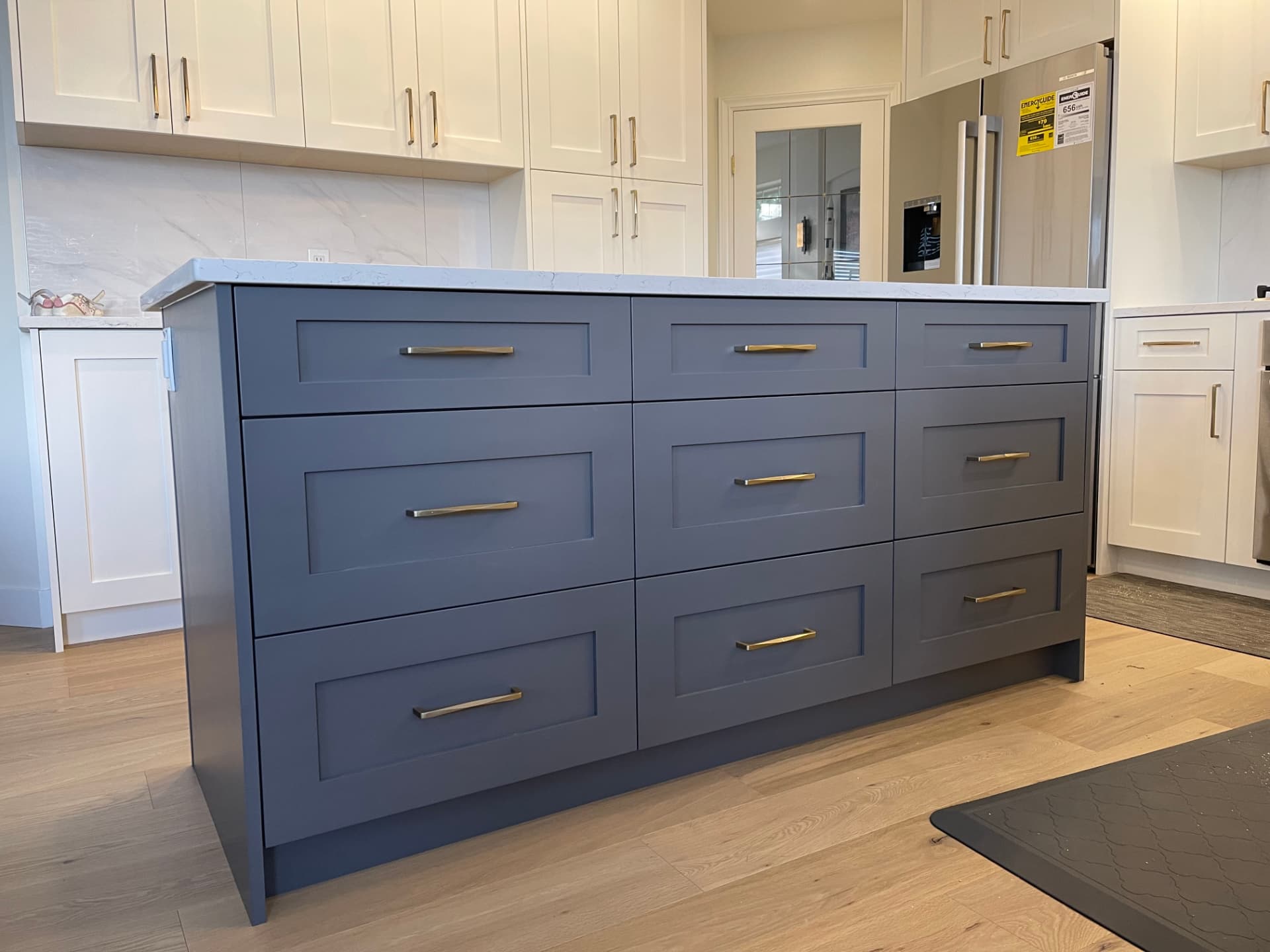 Blue-grey painted kitchen island with white marble countertop, brass counter supports and hardware, white perimeter cabinets, marble range hood, and brass range