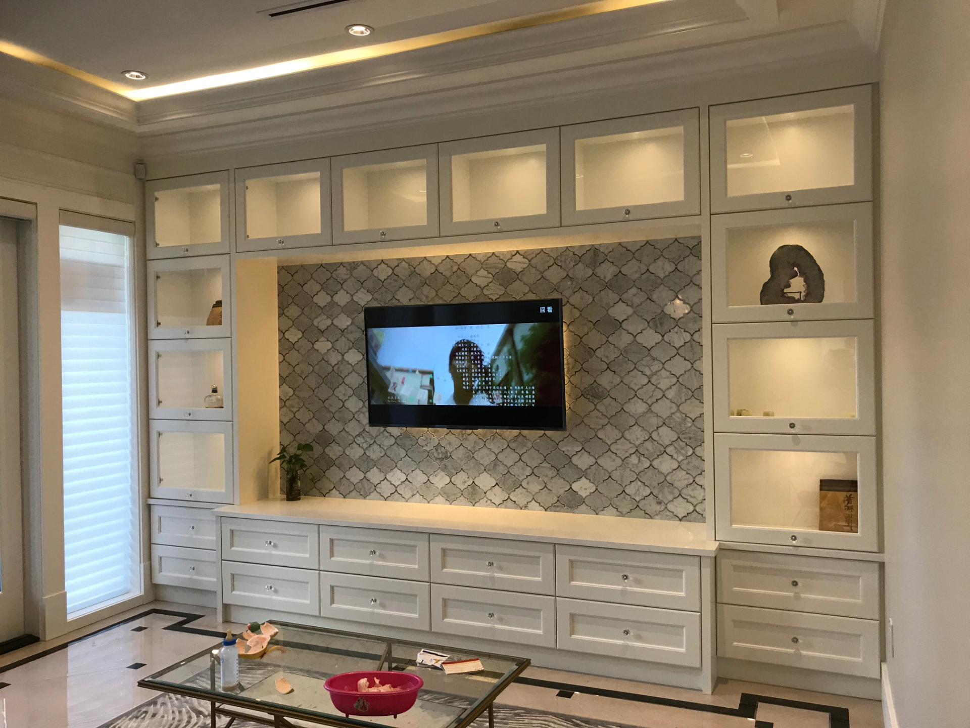 Custom white built-in wall cabinet unit with illuminated glass-front display cabinets, drawer base units, and a wall-mounted television against a backlit grey-and-white arabesque tile panel