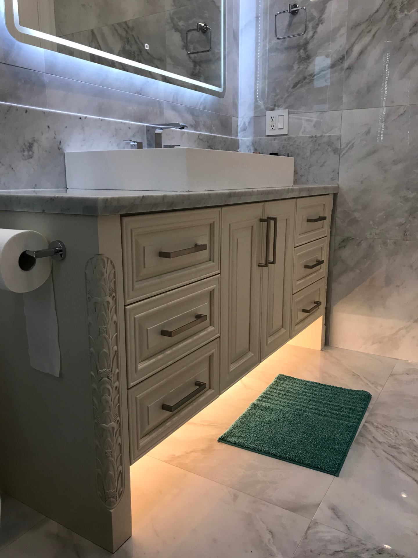 Cream custom bathroom vanity with carved corner detail, marble counter and walls, and backlit mirror