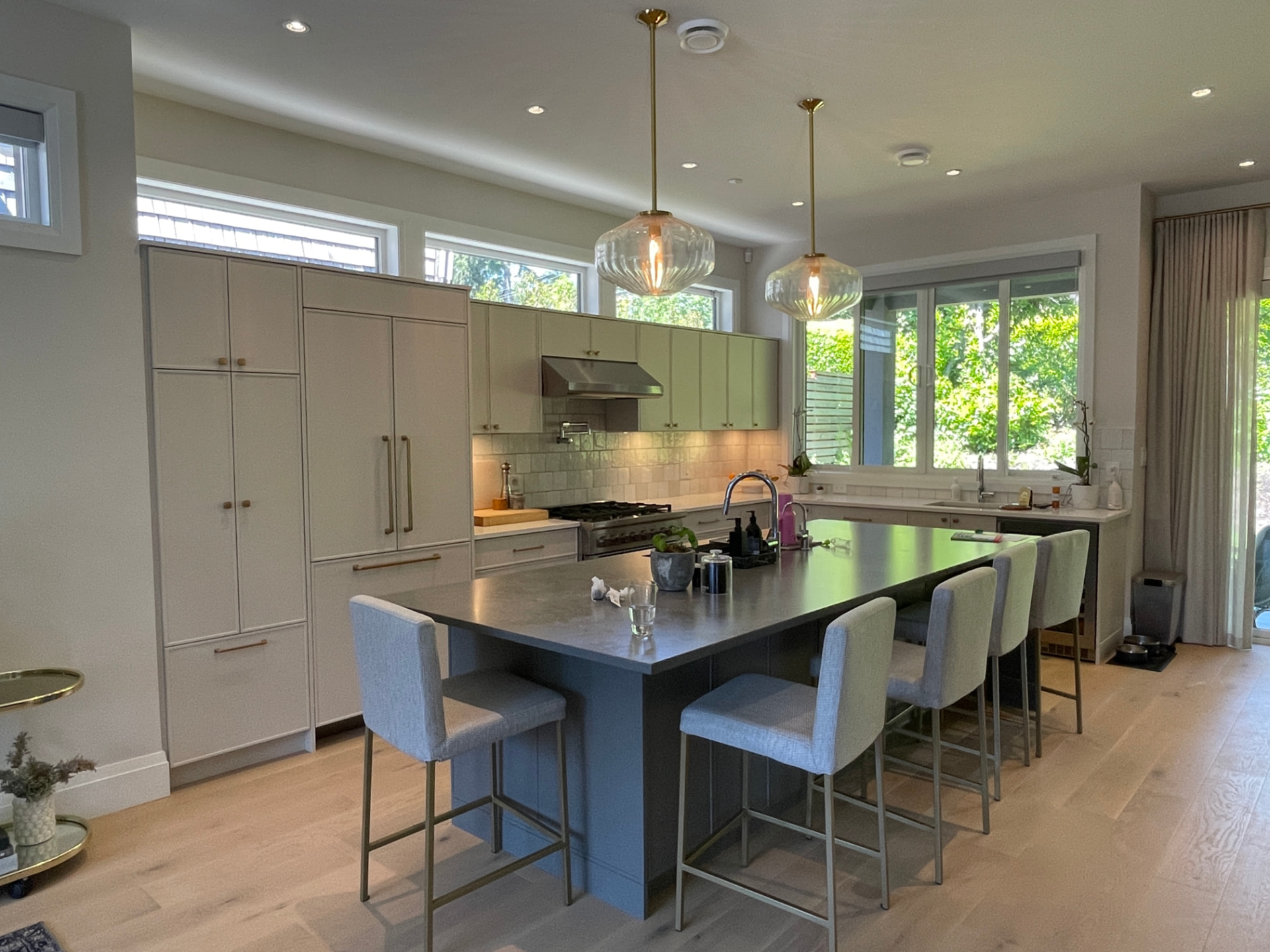 Two-tone kitchen with off-white tall and base cabinets, pale sage upper cabinets, and a slate blue-grey island with a dark matte top, brushed brass bar pulls and round knobs, white quartz perimeter counters, brass-framed counter stools, and fluted glass pendants