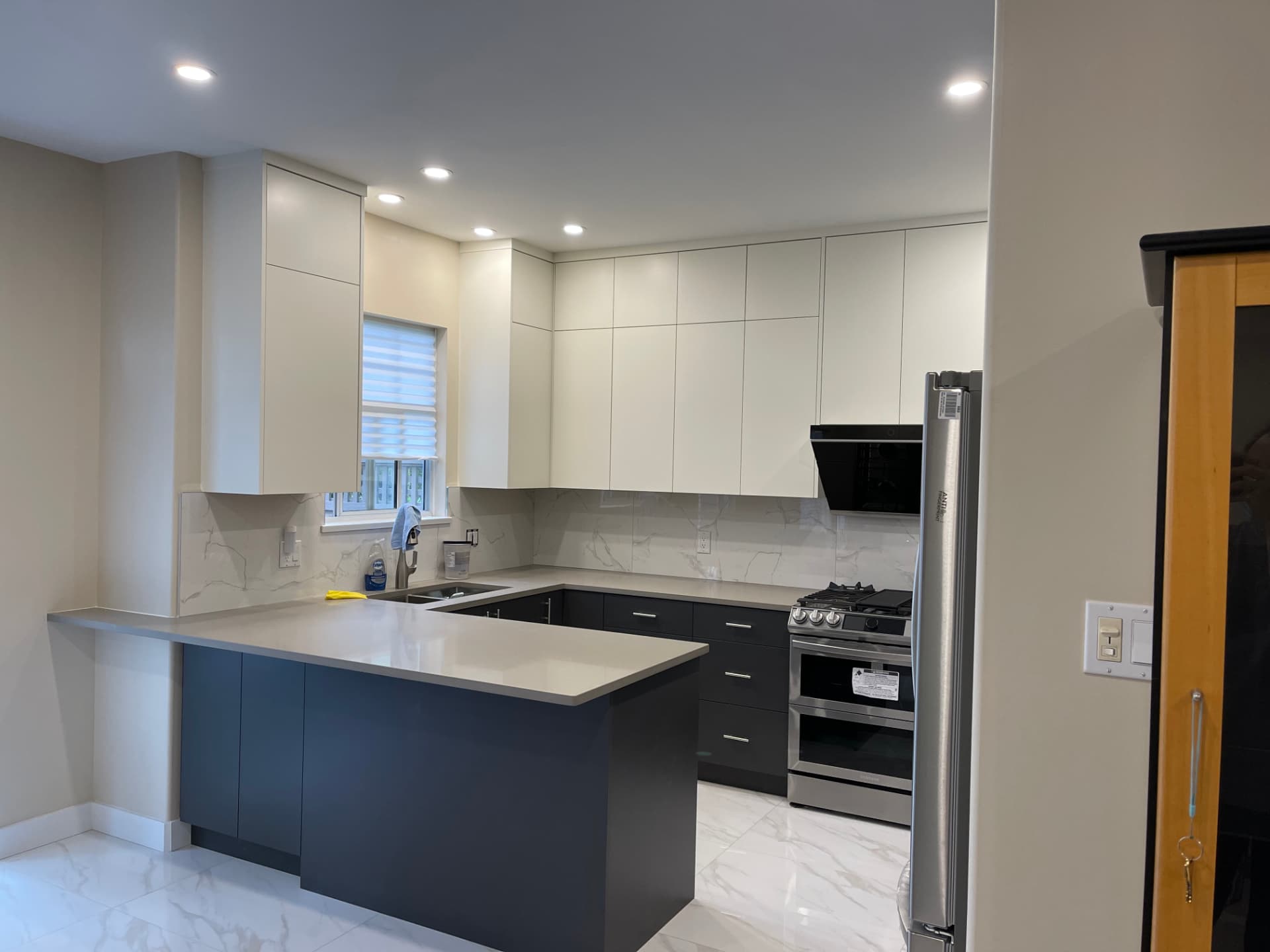 Two-tone kitchen with matte charcoal grey base cabinets and peninsula, light grey flat-panel upper cabinets, marble-veined quartz counters and backsplash, black angled range hood, and stainless steel appliances