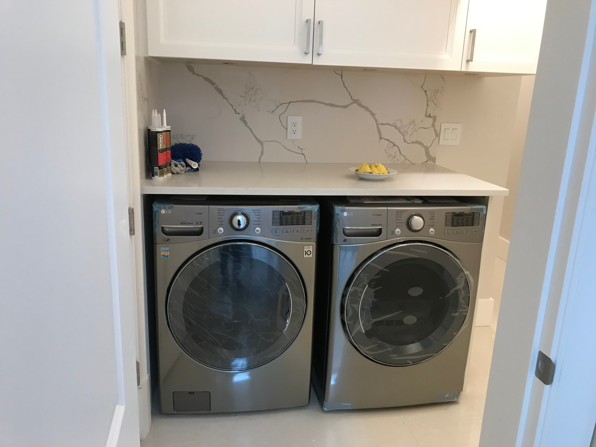 White recessed-panel laundry cabinets with crown molding and brushed bar pulls, white marble-veined quartz counters and a full-height slab backsplash, over a graphite front-load washer and dryer