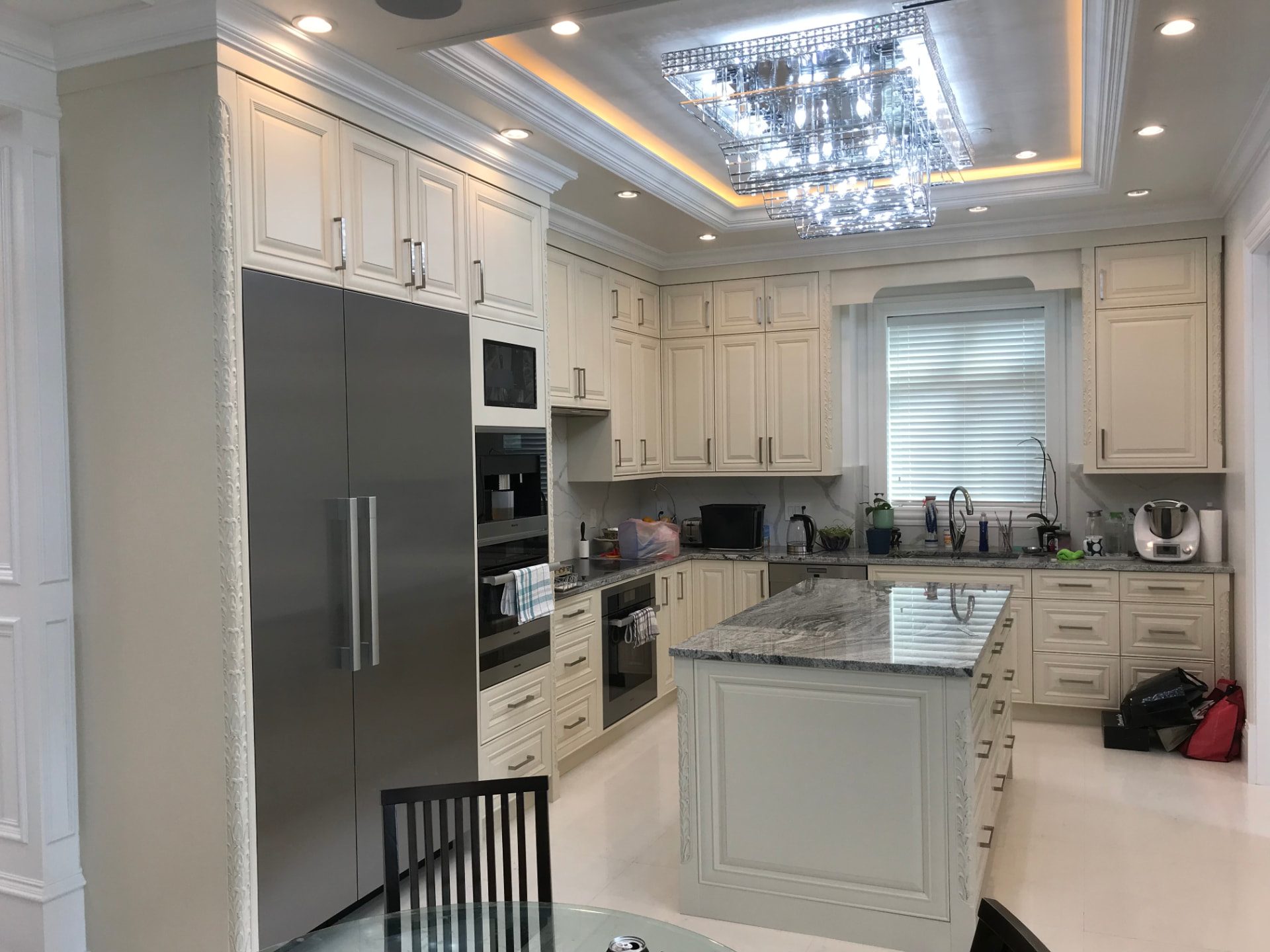Off-white raised-panel kitchen with carved corner trim and brushed metal pulls, grey swirling granite island and perimeter counters, white marble-veined slab backsplash, built-in stainless appliances, and a tiered crystal chandelier in a tray ceiling
