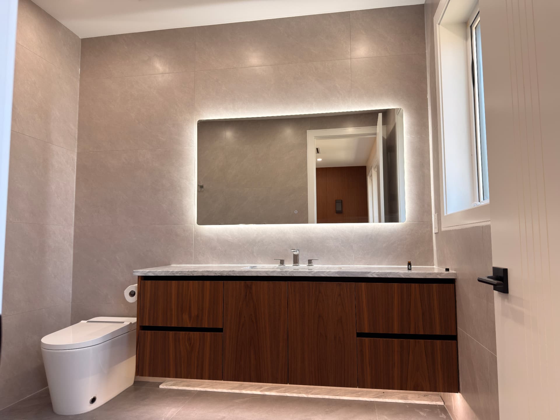 Floating walnut bathroom vanity with a white marble counter and backlit mirror