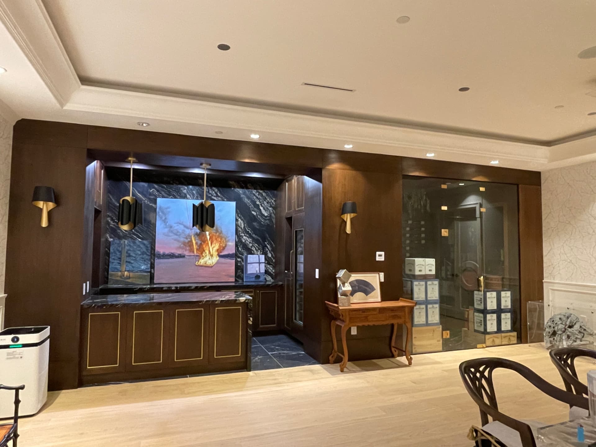 Dark walnut wet bar with black-and-gold veined marble waterfall counters and brass trim, brass-and-black pendant lights, and an adjacent glass-enclosed wine room with backlit bottle walls, a wooden display barrel, and a crystal chandelier