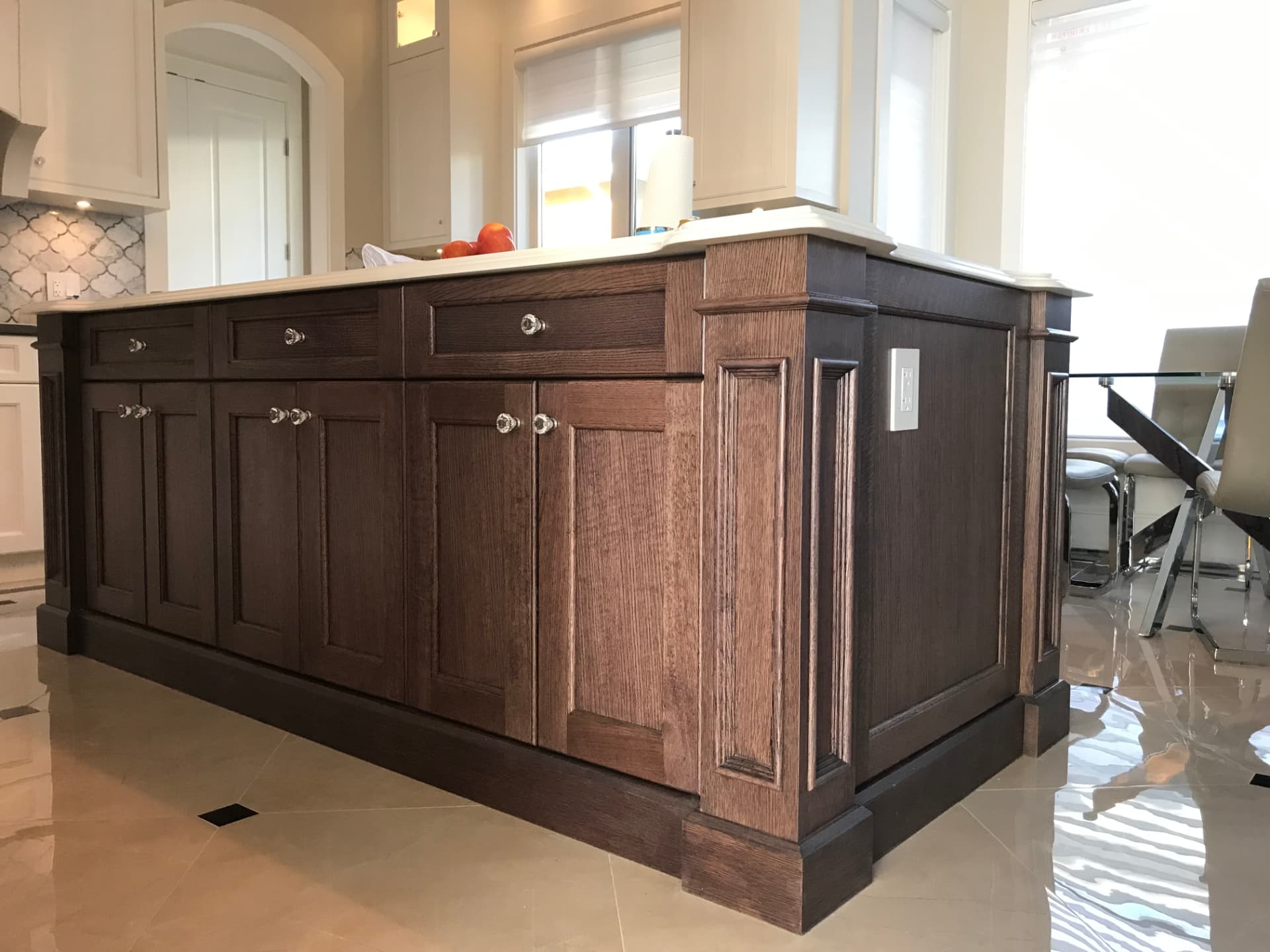 Dark stained-wood kitchen island with white stone countertop and crystal knobs, off-white painted perimeter cabinets, arabesque tile backsplash, and crystal chandelier